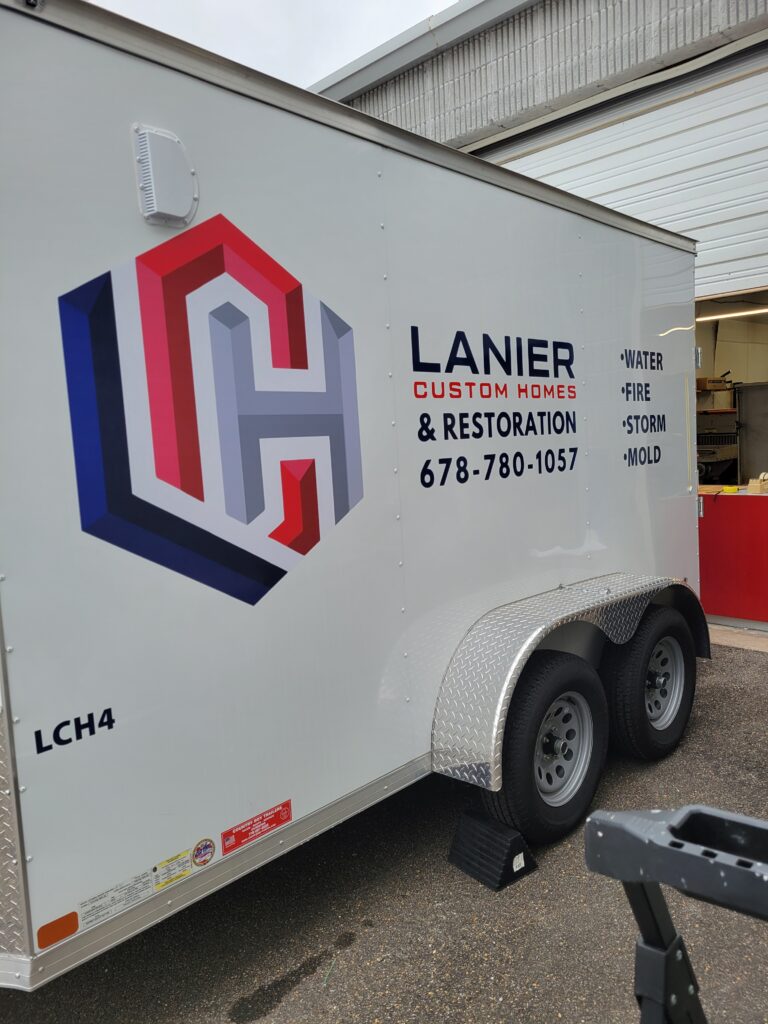 Angled corner view of a white Lanier Custom Homes enclosed cargo trailer showing the seamless application of large vinyl logo sheets near diamond plate rock guards.