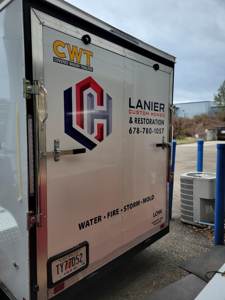 Rear cargo doors of a white Lanier Custom Homes and Restoration enclosed trailer showing a large geometric logo, company phone number 678-780-1057, and a Georgia license plate TY77D52.