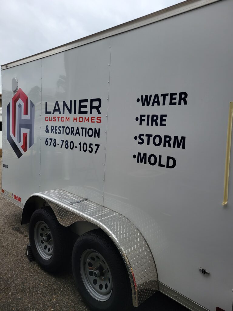 Rear side angle of an enclosed white service trailer displaying bold black vinyl lettering for water, fire, storm, and mold restoration.