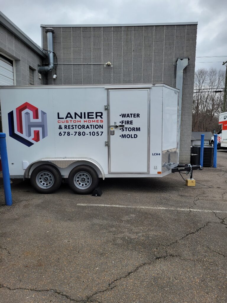 Side view of an enclosed white mobile utility trailer displaying Lanier Custom Homes and Restoration corporate graphics and service listings parked outside a commercial brick building.