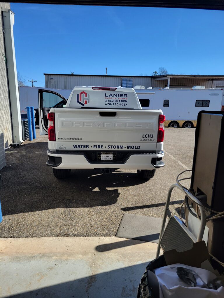 Rear view of a white Chevy Silverado pickup truck tailgate featuring a small corporate logo, unit number LCH3, and lower bumper vinyl text.