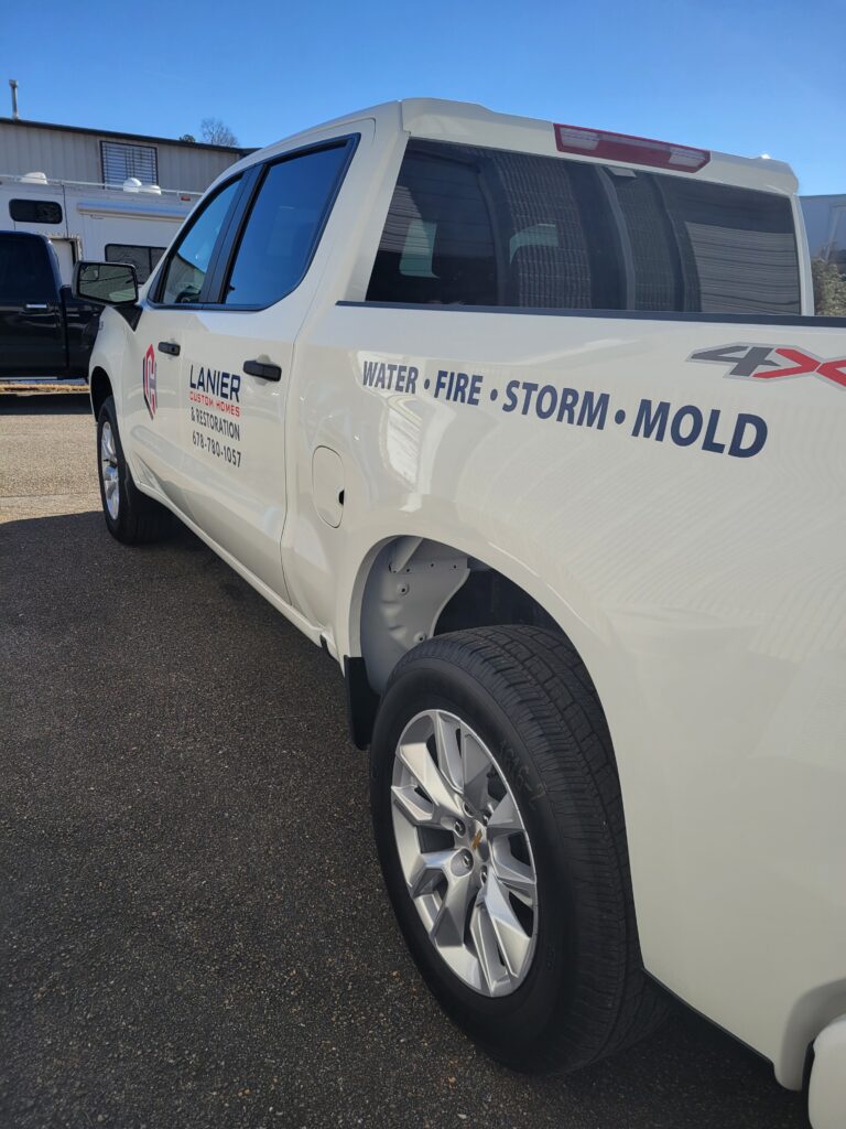 Angled rear-side view of a white commercial truck bed highlighting clean navy blue vinyl text spelling out water, fire, storm, and mold restoration.