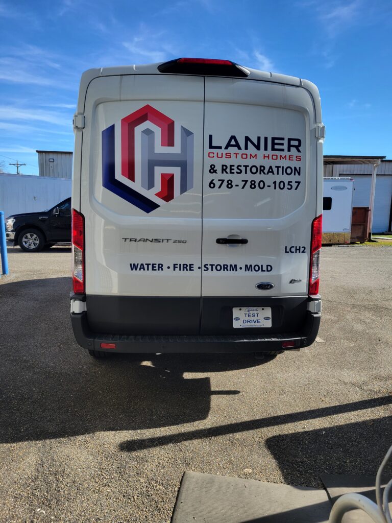Rear view of a white Ford Transit 250 cargo van showing a large interlocking logo, phone number, and lower bumper text for water, fire, storm, and mold.