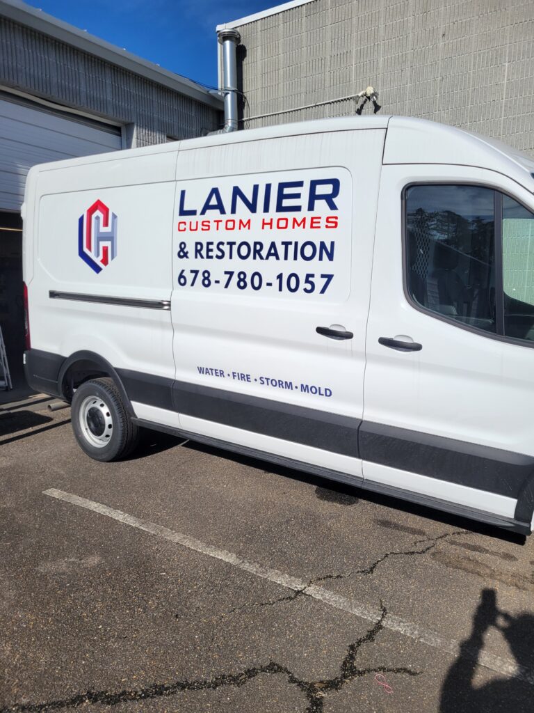 Side profile of a white Ford Transit 250 commercial service van parked outdoors, featuring clean teal, red, and navy blue vinyl fleet decals for Lanier Custom Homes.